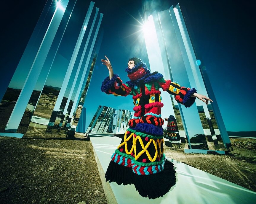 Fashion model in a colorful, textured coat with mirrors in a desert setting, under a bright blue sky and a strong sun.