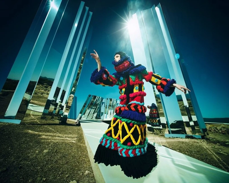 Fashion model in a colorful, textured coat with mirrors in a desert setting, under a bright blue sky and a strong sun.