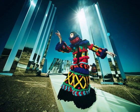 Fashion model in a colorful, textured coat with mirrors in a desert setting, under a bright blue sky and a strong sun.