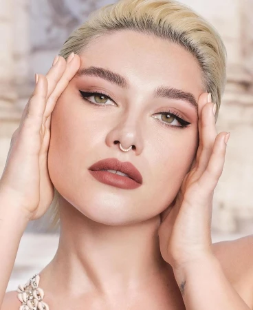 Close-up shot of a fair-skinned adult woman with blonde hair, green eyes, and light pink lipstick posing with hands near her face.