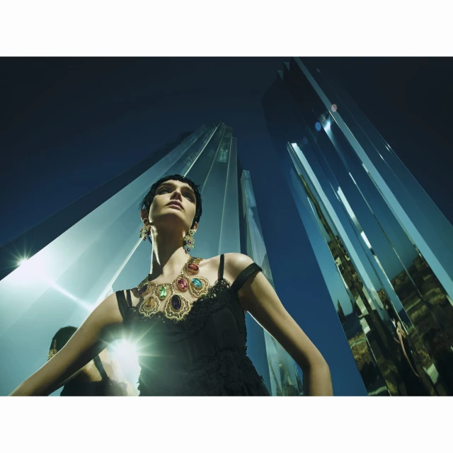 Woman with black dress and ornate necklace stands beneath modern mirrored skyscrapers in a low-angle fashion or jewelry campaign shot.