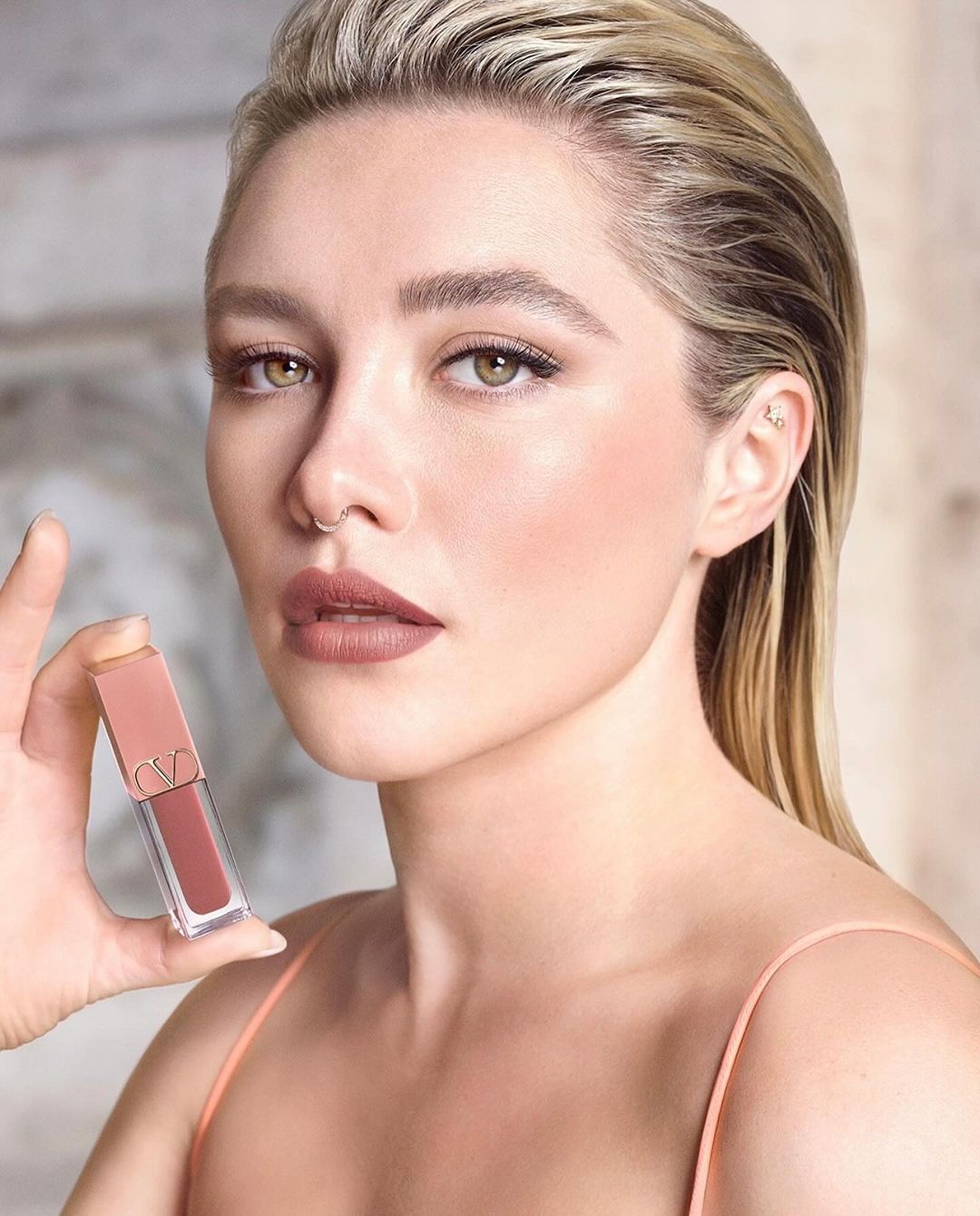 Close-up of an adult female holding a tube of lipstick, showcasing makeup and blonde hair, set against a neutral-toned backdrop.