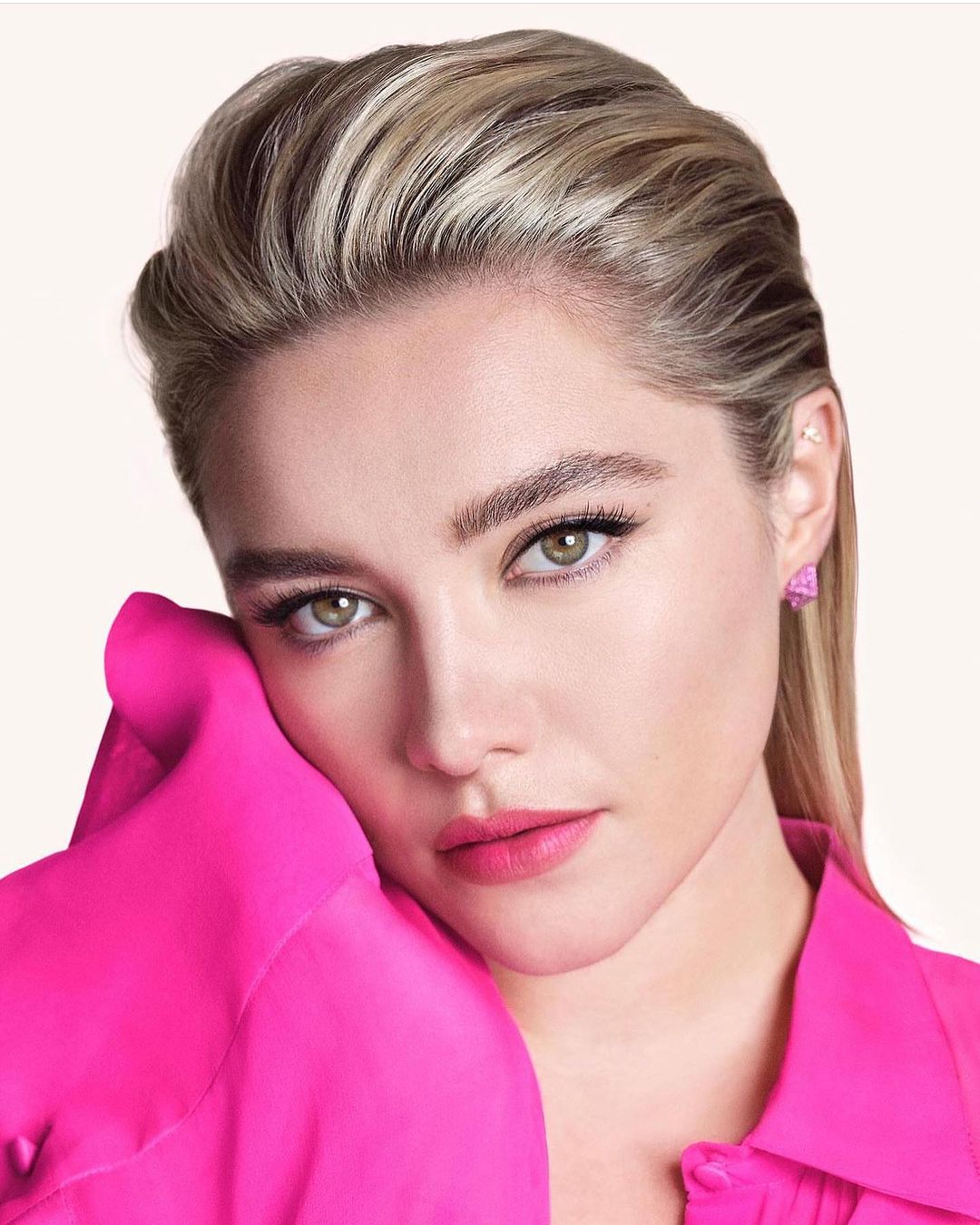 Close-up portrait of actress Florence Pugh with slicked-back blonde hair wearing a bright pink buttoned shirt.