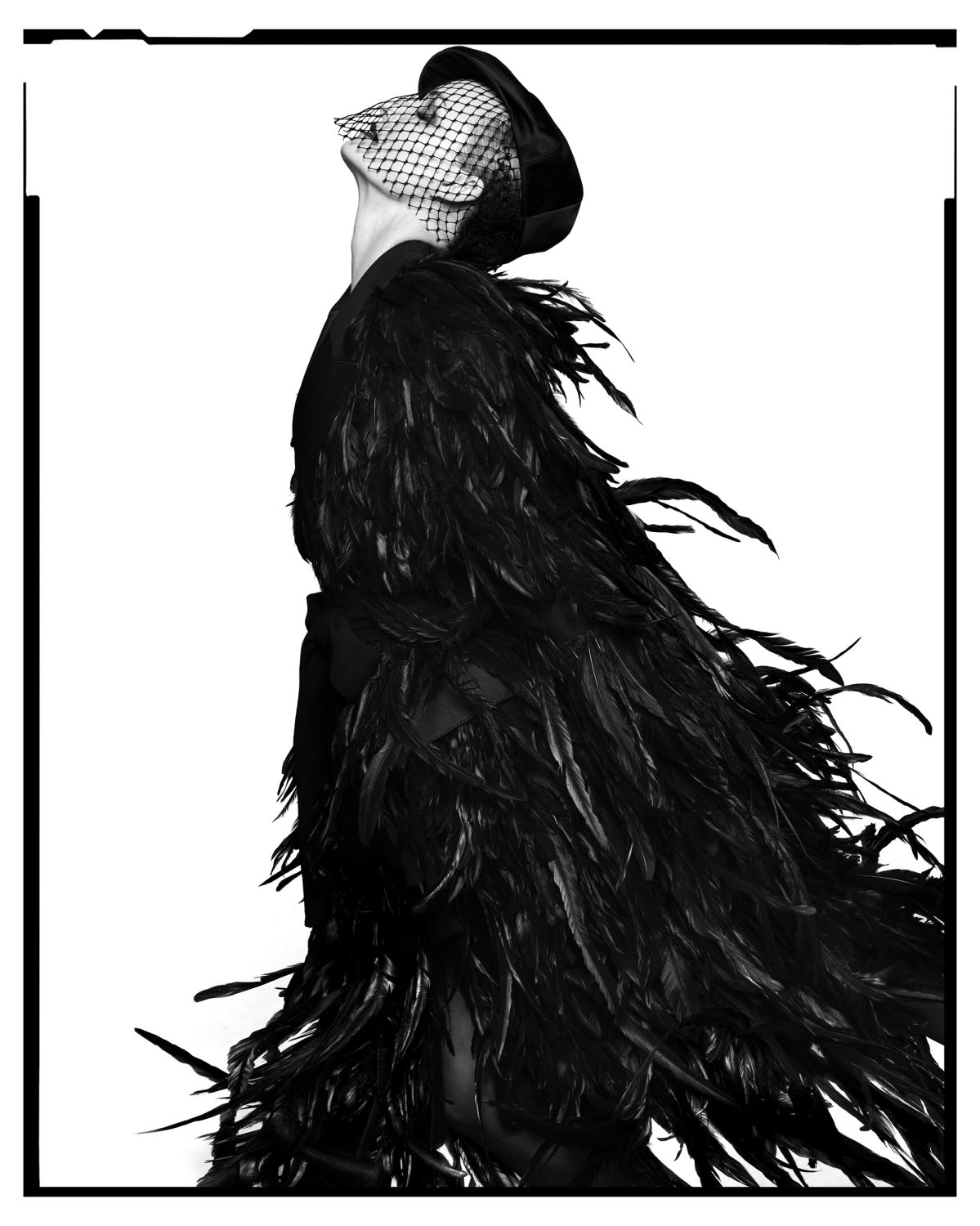 Black and white fashion portrait of a woman in feather coat, net veil, and hat looking up against a white background, framed by a black border.