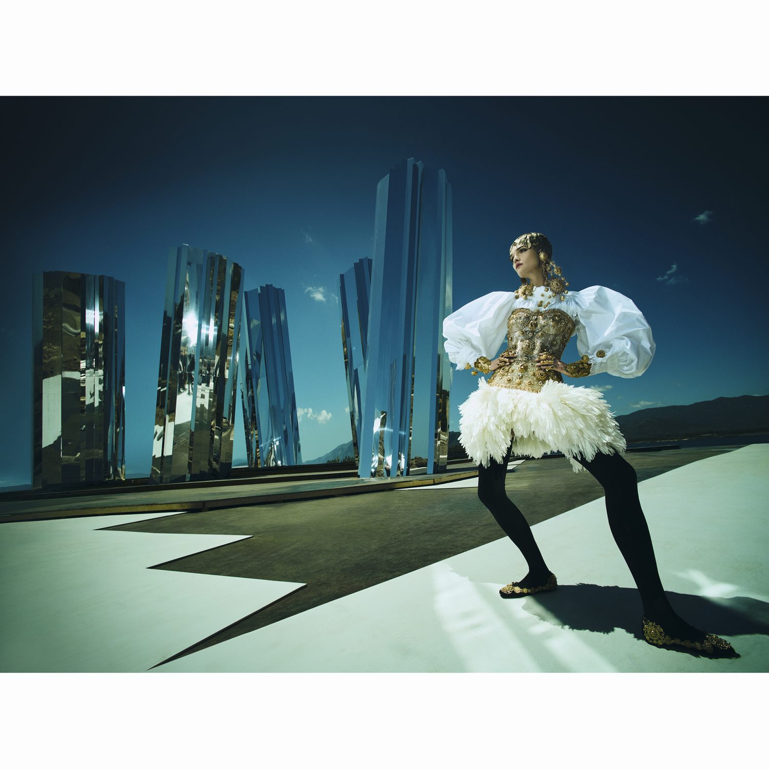 Fashion model posing in a shimmering gold and white dress with a unique architectural backdrop and a clear blue sky in an outdoor setting.