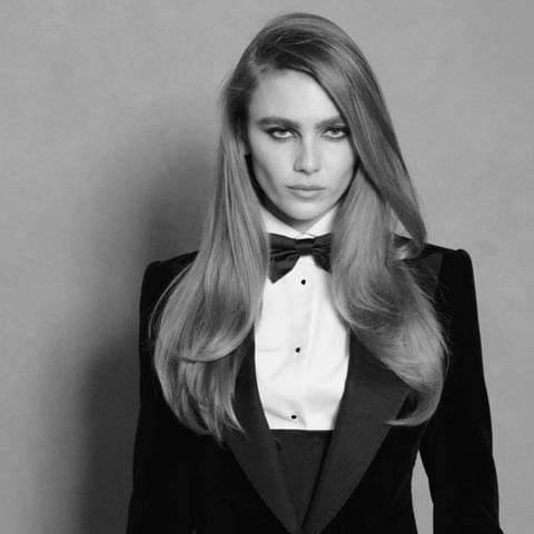 Black and white portrait of a young adult female with long hair wearing a tuxedo with a bow tie against a neutral gray background.