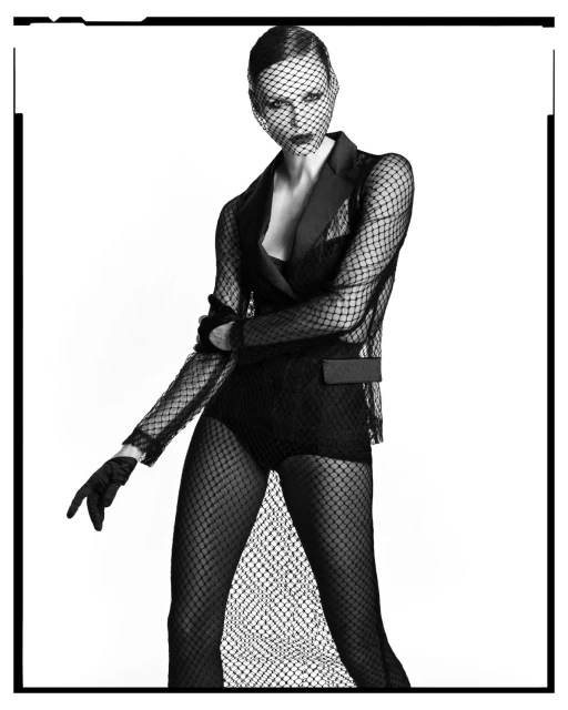 Black-and-white portrait of a woman wearing a mesh veil and blazer-style jacket, in a studio with a white backdrop.