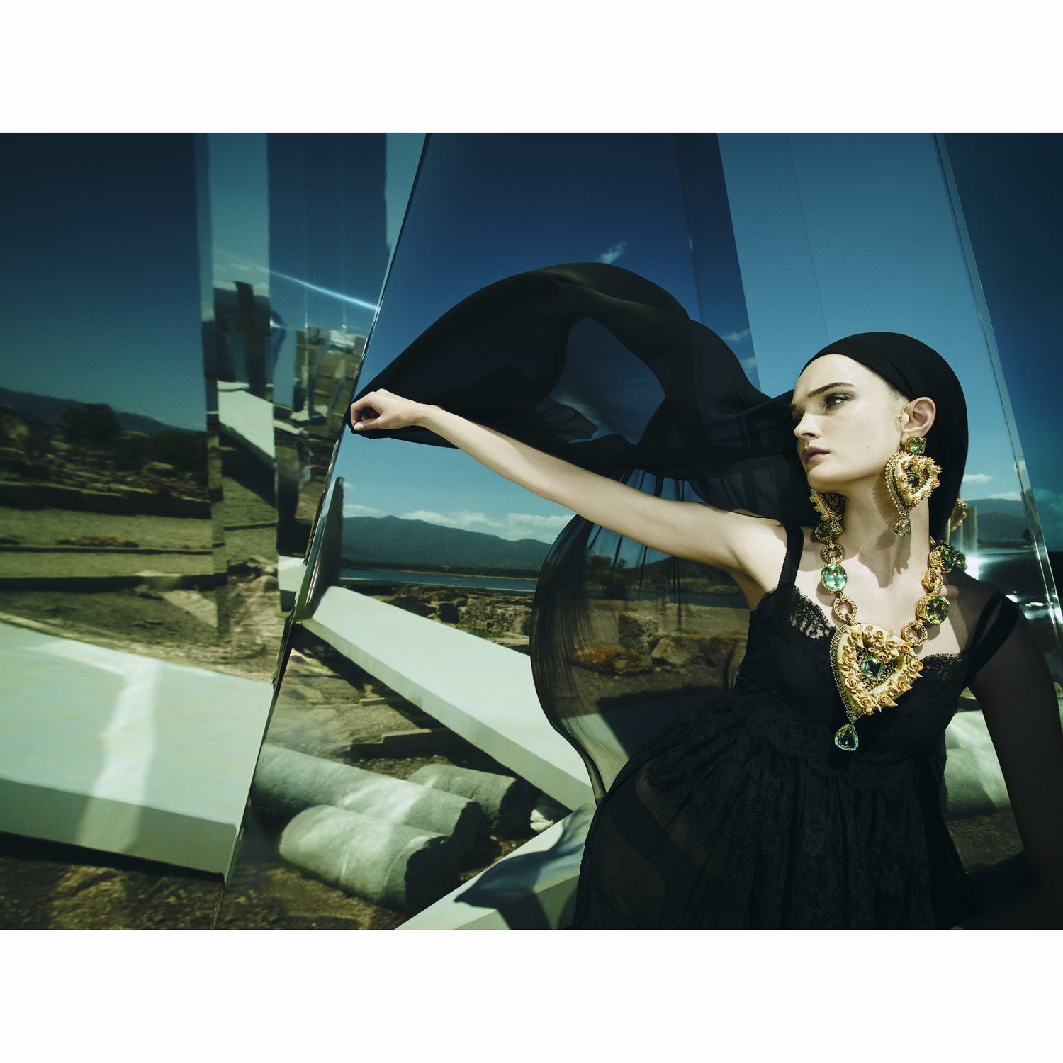 Fashion shot of a woman wearing a black dress, gold jewelry, and a headscarf, posing in front of reflecting mirror-like structures.