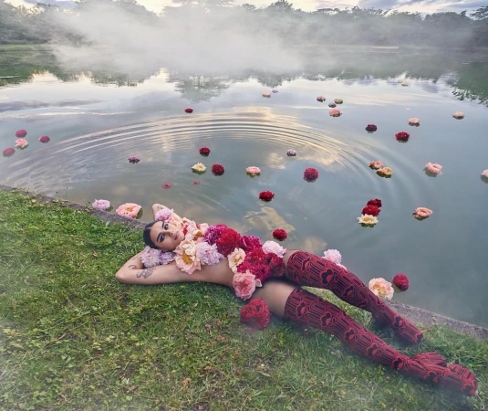 Woman lying on grassy bank beside water covered in scattered flower blossoms with mist floating over the surface