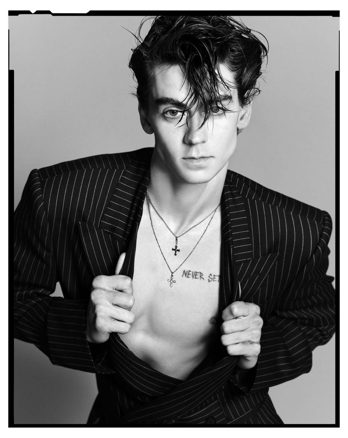 Black and white portrait of a young adult male model in a pinstripe blazer, jewelry, chest tattoo, and slicked-back hair, studio lighting.
