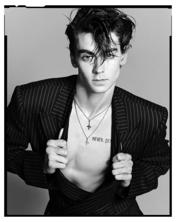 Black and white portrait of a young adult male model in a pinstripe blazer, jewelry, chest tattoo, and slicked-back hair, studio lighting.