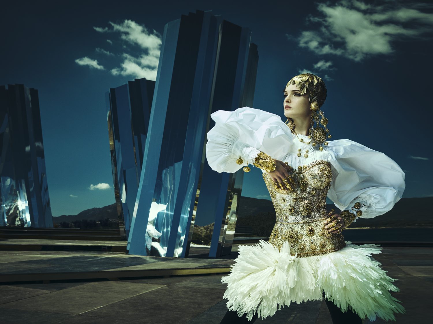 Woman in an elaborate white and gold costume poses in front of mirror-like sculptures and a dark blue sky with scattered clouds.