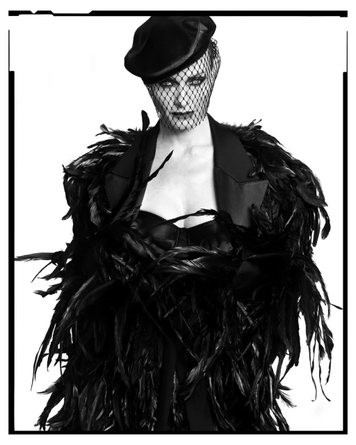 Woman wearing a black feathered jacket and a veil over her face, posing in a close-up shot against a bright white background.