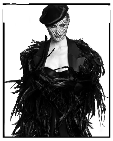 Woman wearing a black feathered jacket and a veil over her face, posing in a close-up shot against a bright white background.