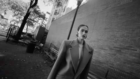 A stylish adult woman in a coat poses in an urban park setting, presented in a black and white photograph.