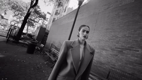 A stylish adult woman in a coat poses in an urban park setting, presented in a black and white photograph.