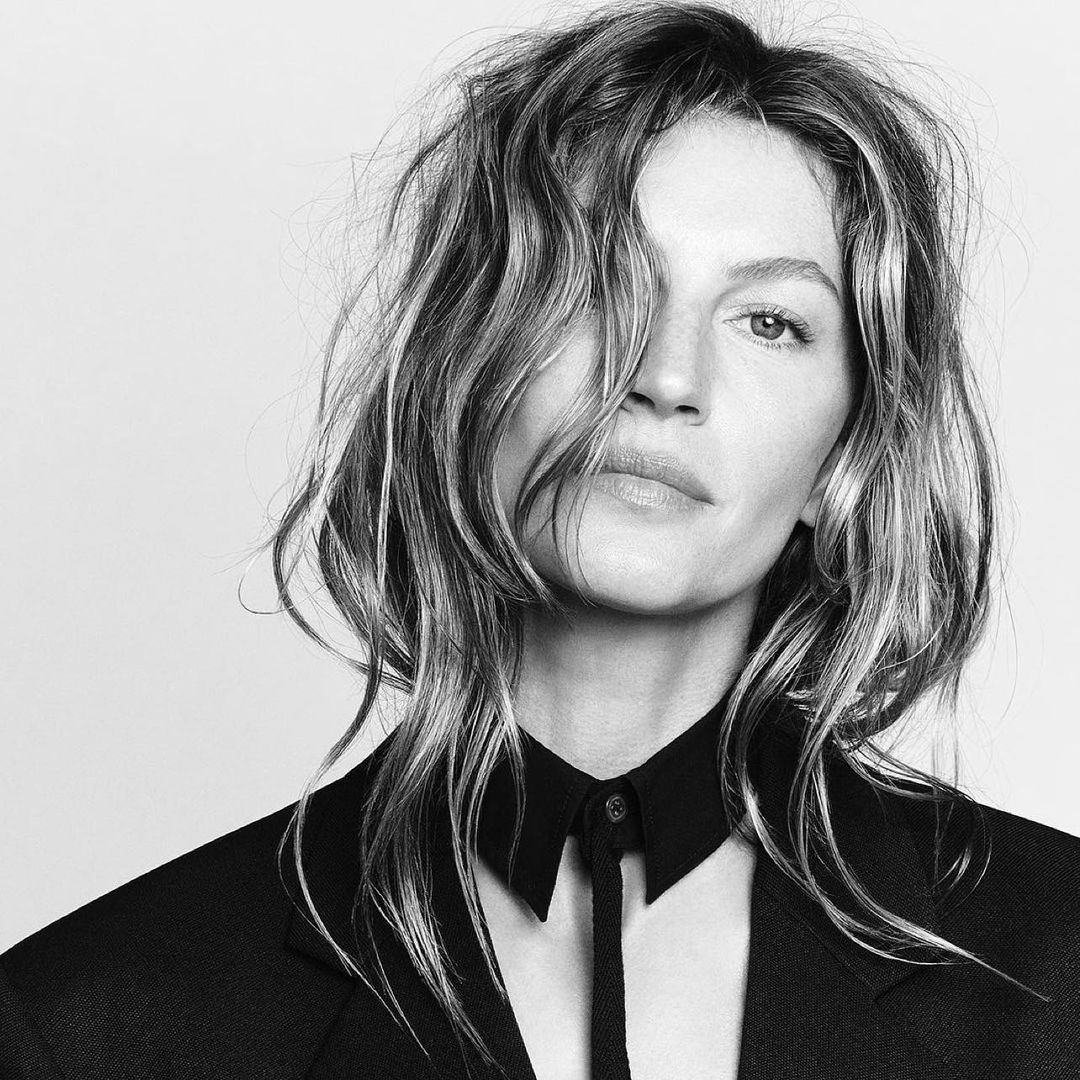 Black and white portrait of a woman with wavy hair wearing a dark blazer and collared shirt against a pale gray background.