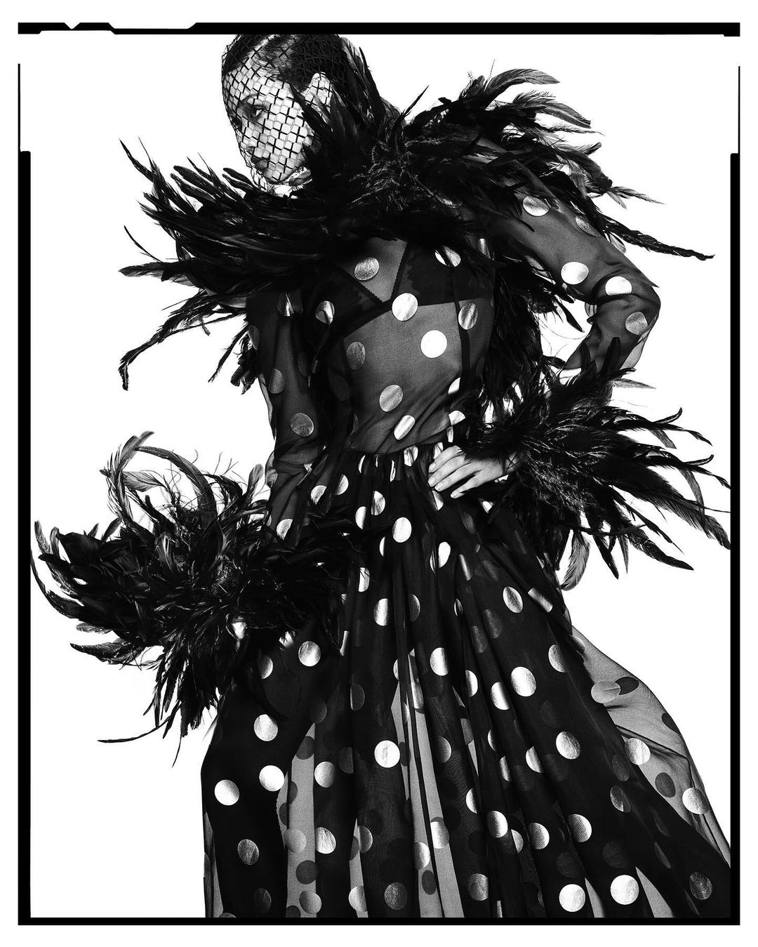 Fashion model wearing black polka dot dress with feathered boa and veiled headpiece against a white background in a studio shot.
