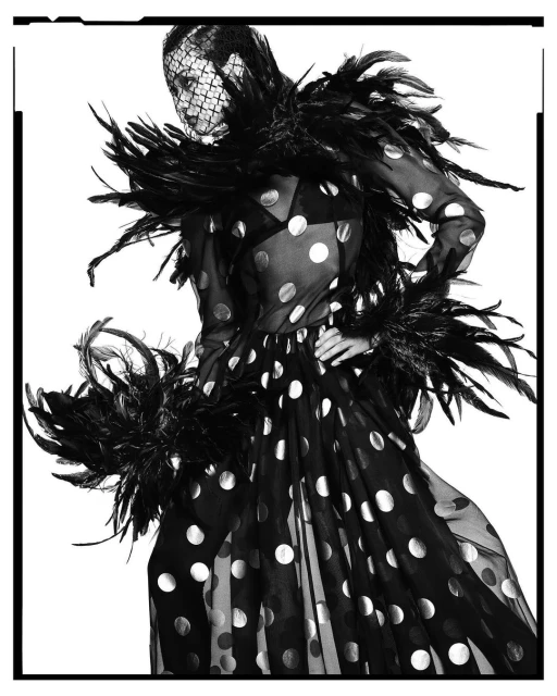 Fashion model wearing black polka dot dress with feathered boa and veiled headpiece against a white background in a studio shot.