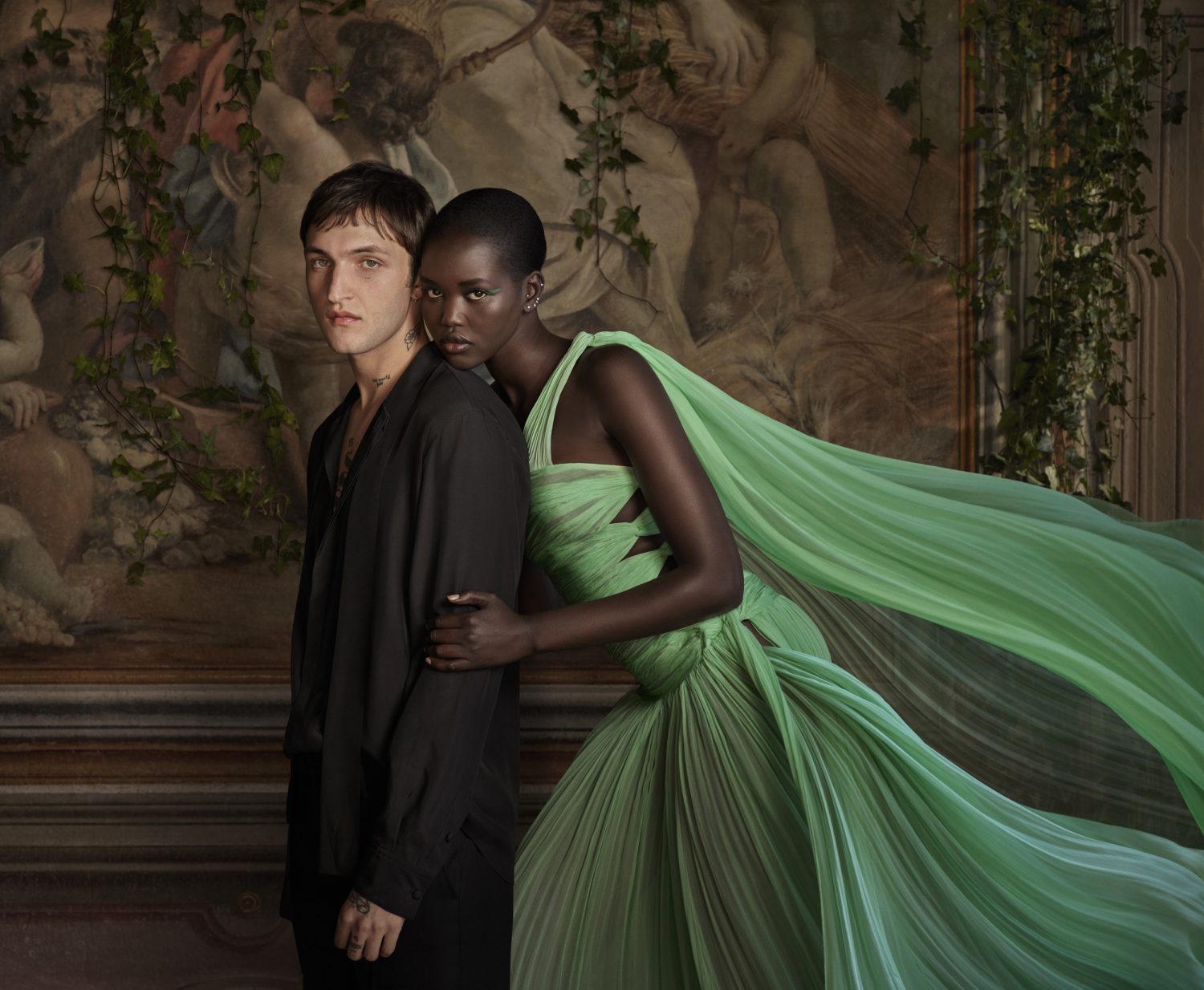 An adult man and woman pose together in a fashionable indoor setting with draped ivy and classical mural in the background.