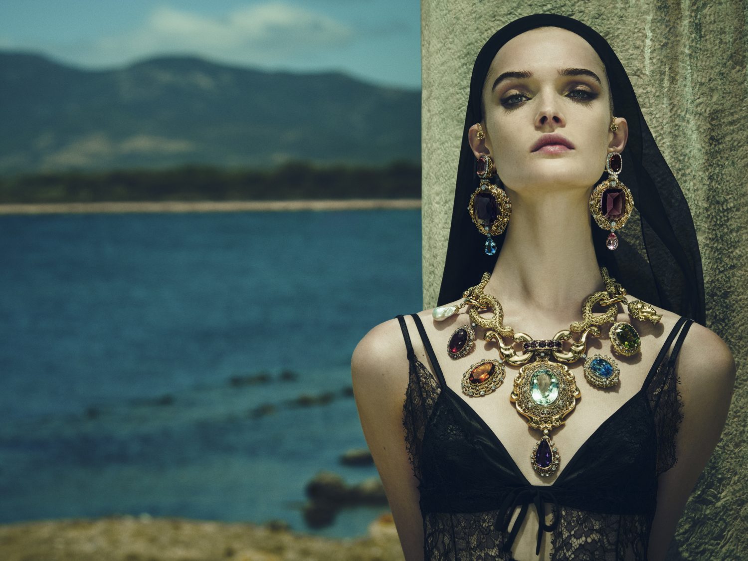 Model wearing ornate jeweled statement necklace and black lace dress against coastal landscape by Luigi and Iango