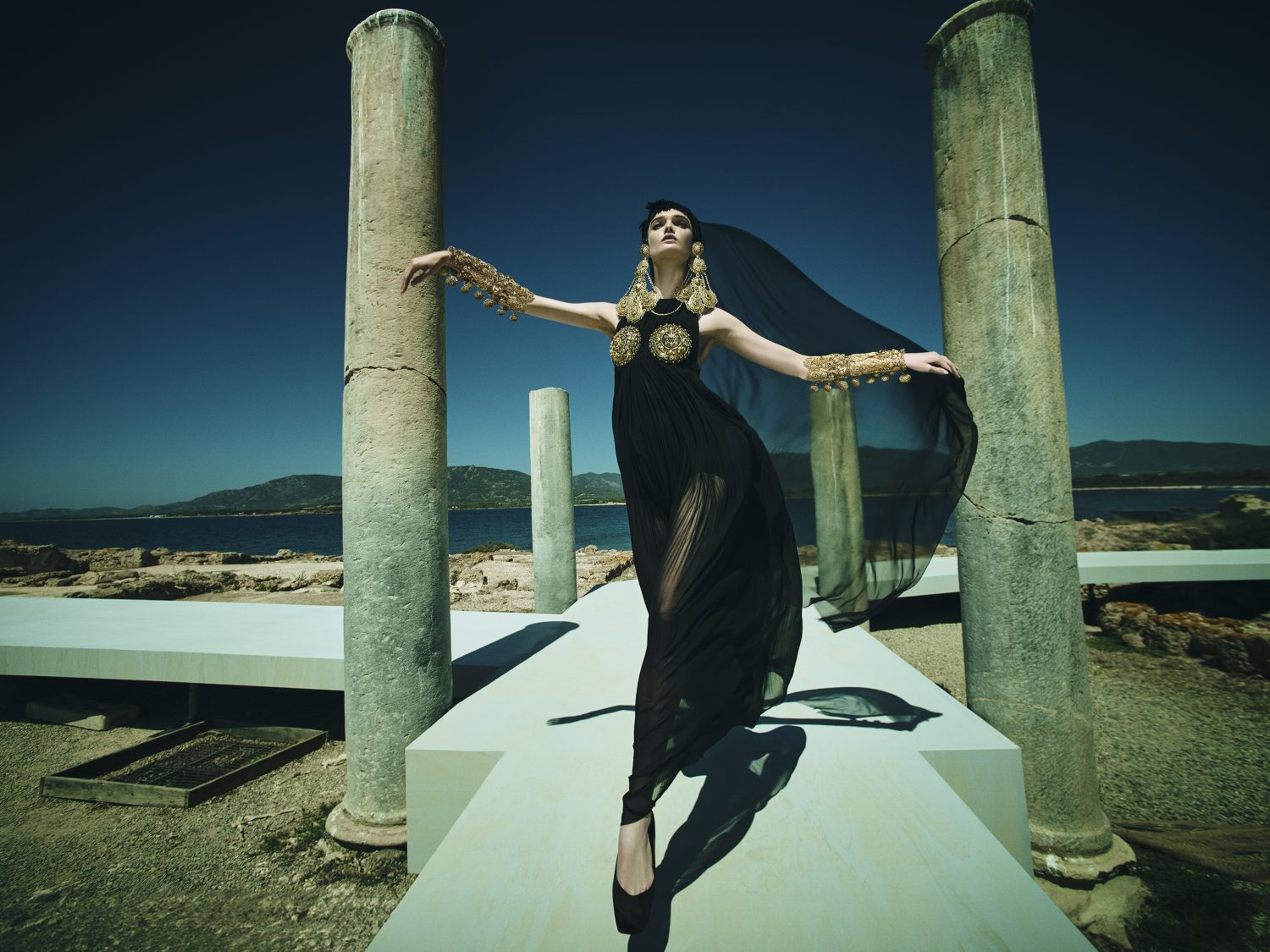 A woman in a black dress with gold jewelry stands on a white platform among stone columns, with the sea and a blue sky in the background.