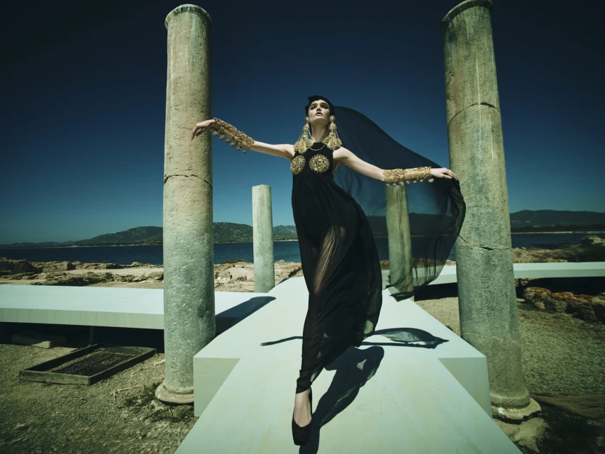 A woman in a black dress with gold jewelry stands on a white platform among stone columns, with the sea and a blue sky in the background.