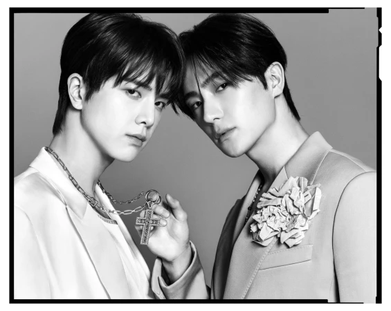 Two men posing closely together in formal blazers with a cross necklace and floral lapel pin in a monochrome portrait.