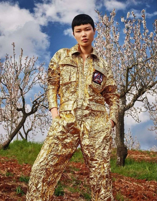 A woman in a gold foil jumpsuit stands in a field with flowering trees and a cloudy sky, showcasing a unique fashion style in nature.