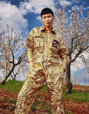 A woman in a gold foil jumpsuit stands in a field with flowering trees and a cloudy sky, showcasing a unique fashion style in nature.
