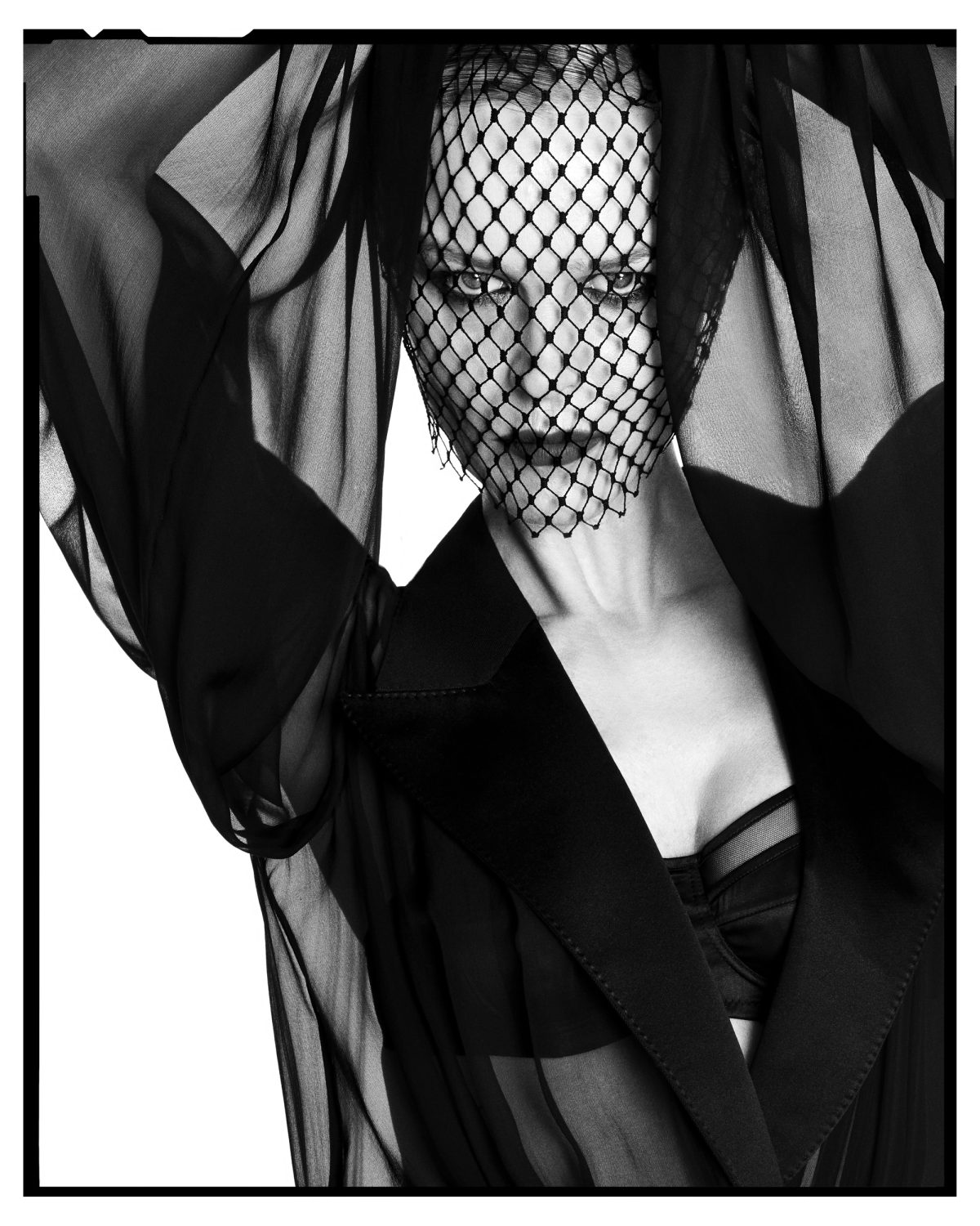 Monochrome portrait of an adult female model wearing a black veil and blazer against a bright white background in a studio setting.