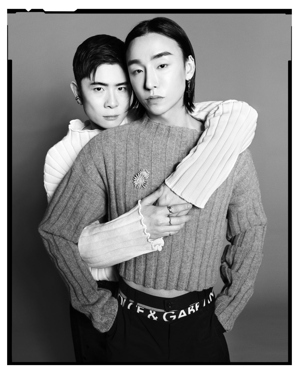 Monochrome portrait of two young adults embracing indoors. One is wearing a grey sweater with a sunflower pin, the other in white.