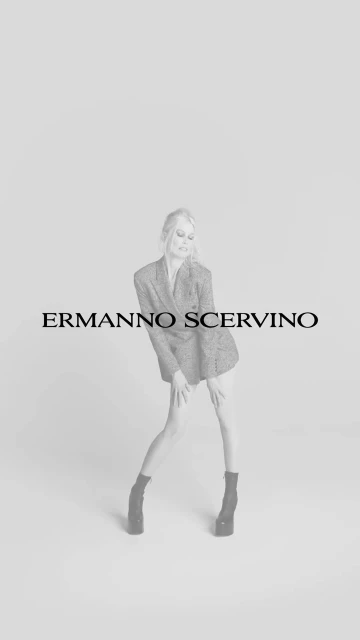 Monochrome fashion shot of a blonde female model in a blazer dress and platform boots, posing in a photo studio with the Ermanno Scervino logo.