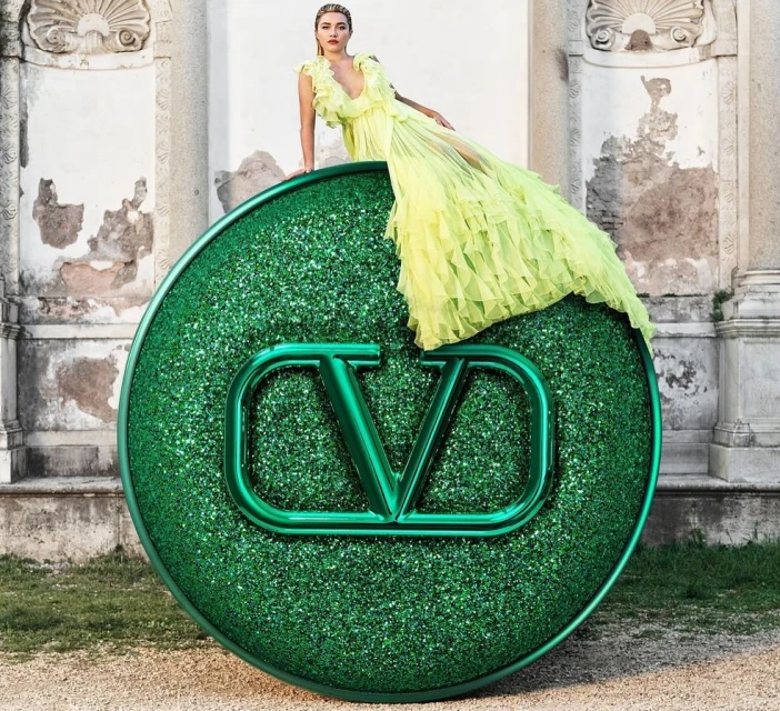 Adult woman in a yellow dress posing on a giant, green Valentino logo sculpture outside during daytime; fashion, beauty, and lifestyle concept.