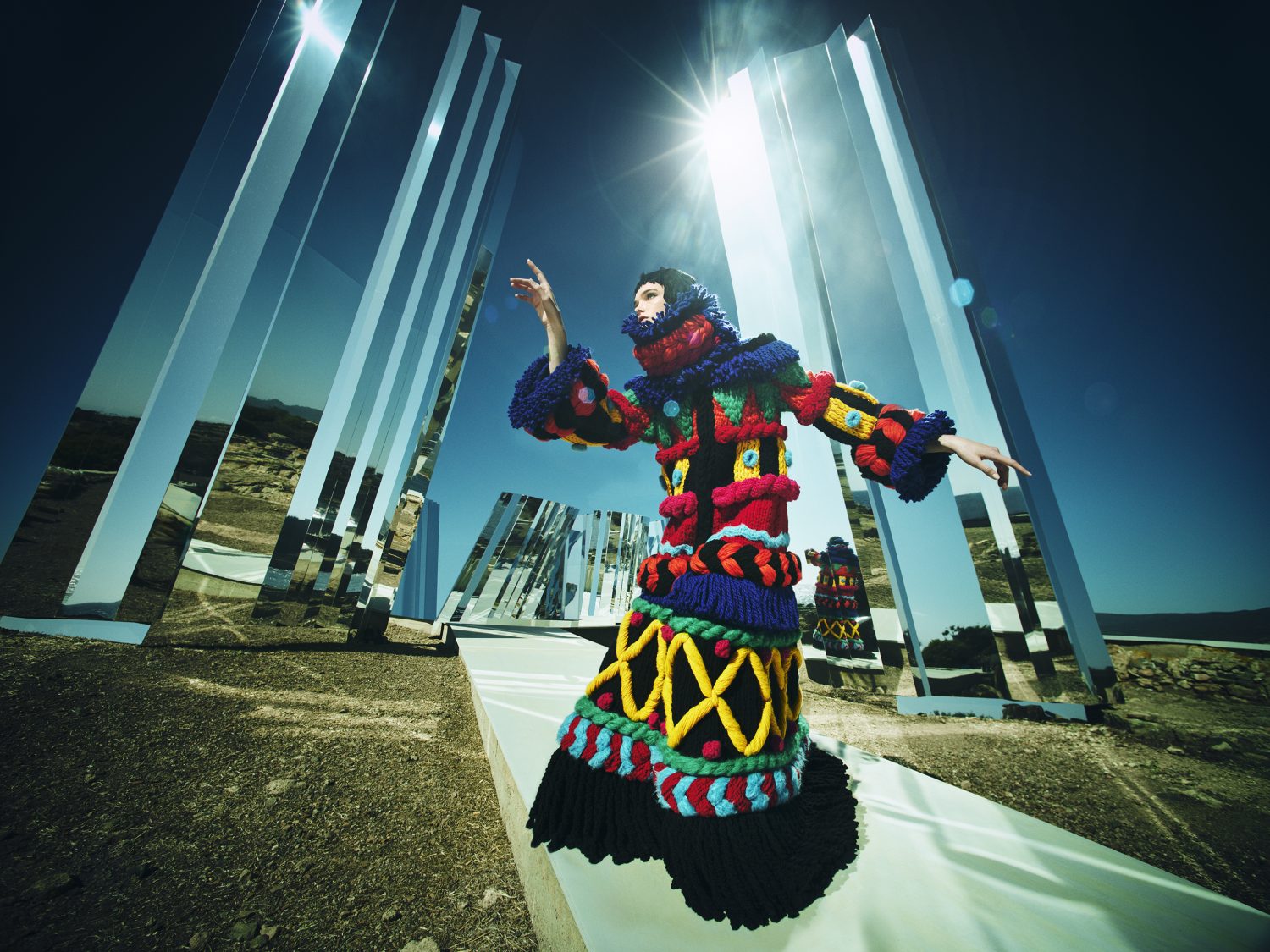 Fashion shot of a woman in colorful knitwear posing in a desert landscape with mirrored monoliths under a clear sky and bright sun.