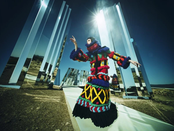 Fashion shot of a woman in colorful knitwear posing in a desert landscape with mirrored monoliths under a clear sky and bright sun.