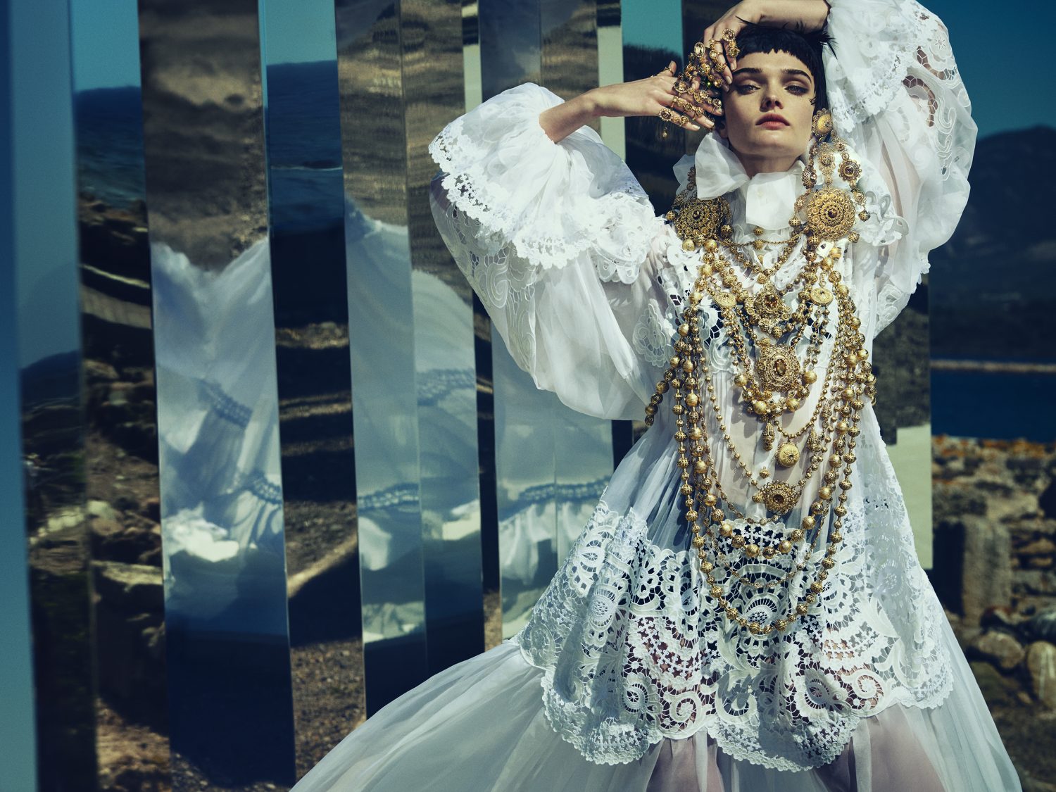 Woman in a white lace dress with gold jewelry poses outdoors, reflected in rectangular mirrors against a natural landscape backdrop.