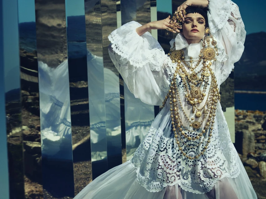 Woman in a white lace dress with gold jewelry poses outdoors, reflected in rectangular mirrors against a natural landscape backdrop.