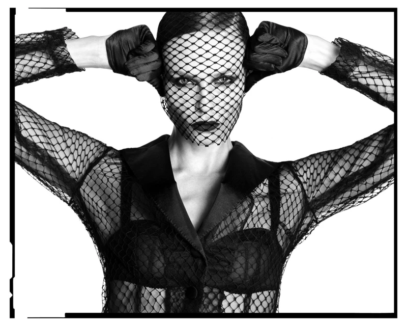 Woman with black net veil and gloves poses in a black jacket against a white background, creating a dramatic fashion portrait.