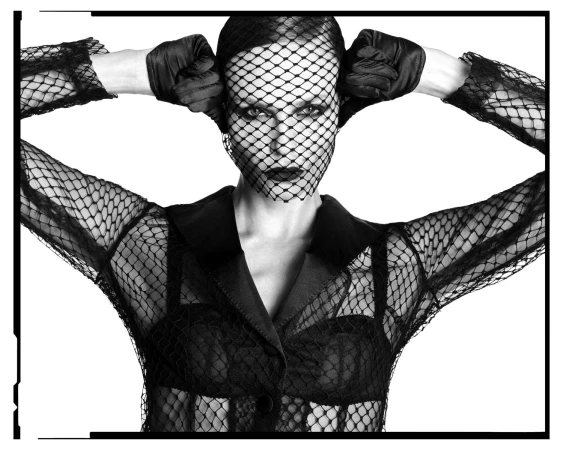 Woman with black net veil and gloves poses in a black jacket against a white background, creating a dramatic fashion portrait.