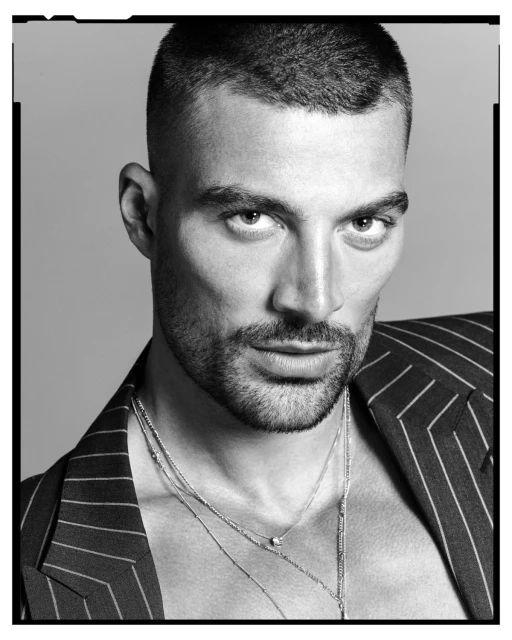Black and white portrait of a male model wearing a pinstripe blazer and multiple necklaces in a photo studio with grey backdrop.