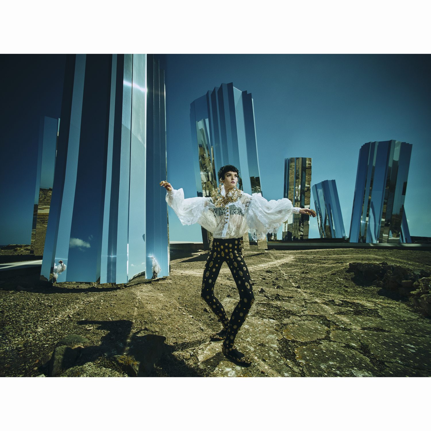 A person in a ruffled white shirt and patterned leggings poses in a desolate landscape with mirrored monoliths under a clear blue sky.