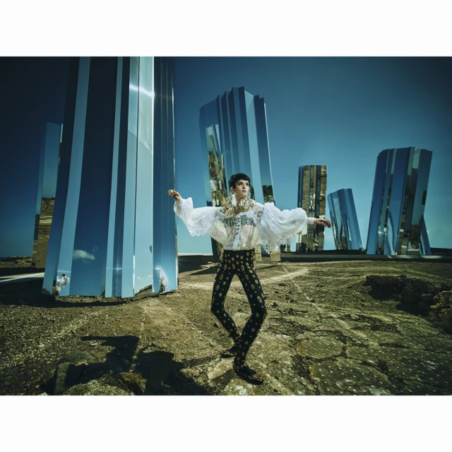 A person in a ruffled white shirt and patterned leggings poses in a desolate landscape with mirrored monoliths under a clear blue sky.