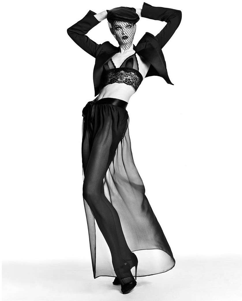 Black and white fashion shot of a woman in lingerie, sheer skirt, jacket, cap, and veil posing against a bright white backdrop.