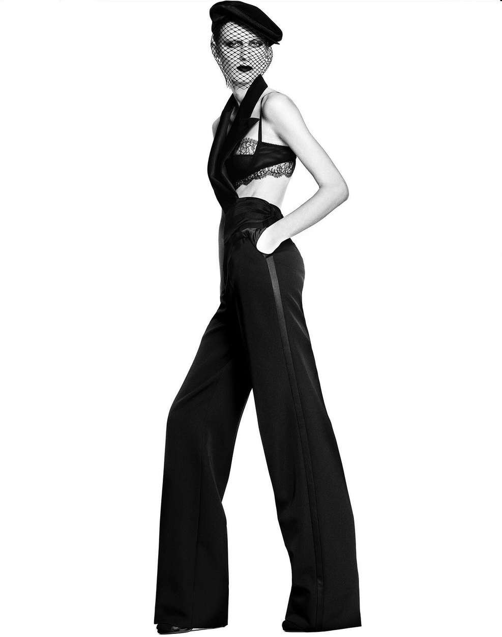 Full-length studio shot of a woman in stylish black lace top, vest, wide-leg trousers, and beret with veil, hands in pockets, black and white.