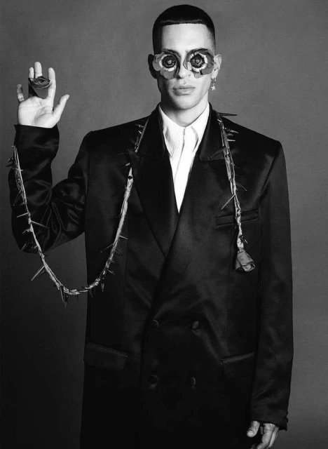 Black and white portrait of a man wearing artistic glasses and a black jacket with spike embellishments, posing indoors.
