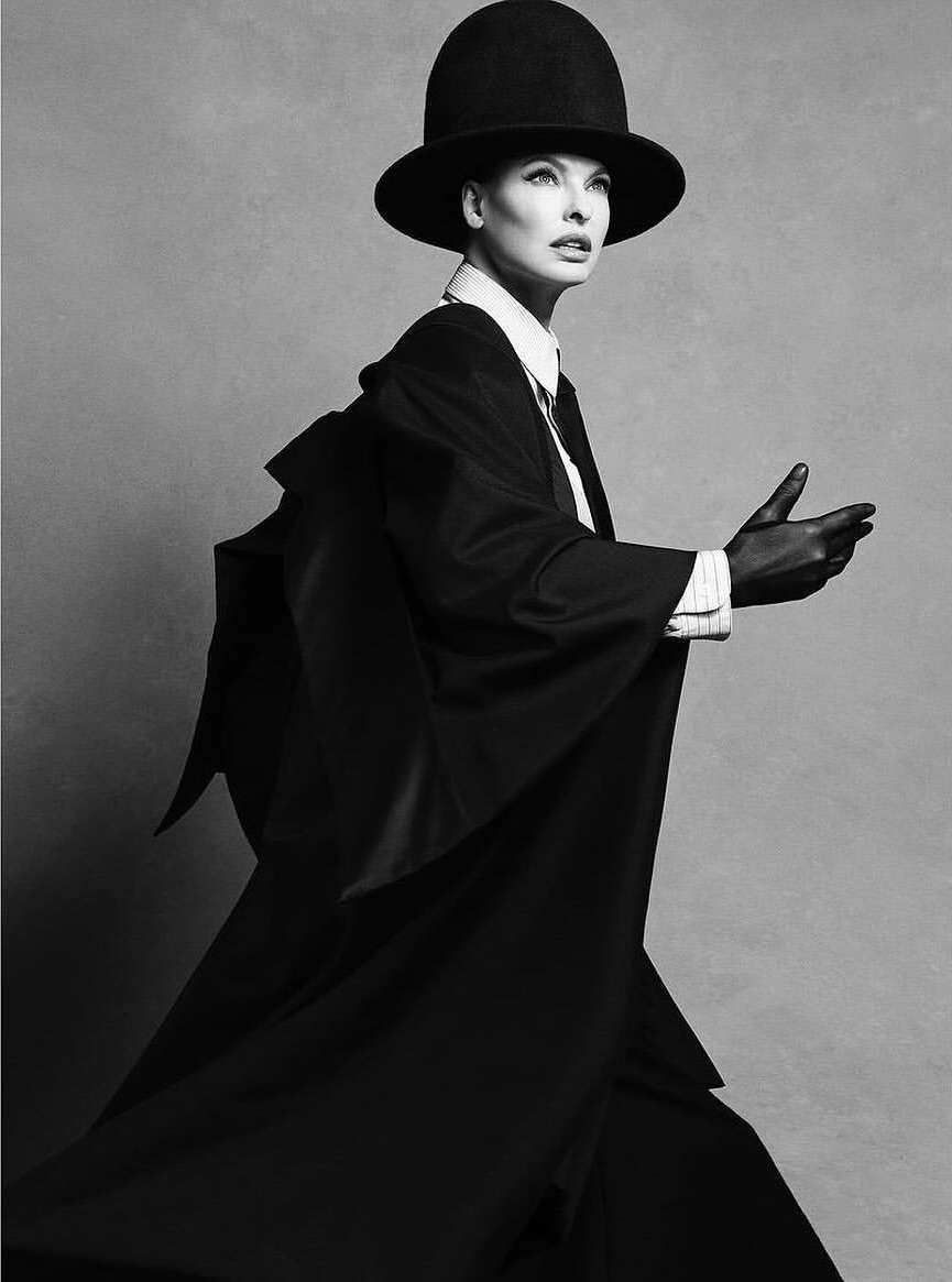 Black and white fashion shot of a model wearing a hat and robe with outstretched hands, photographed in a photo studio.