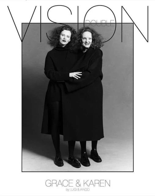 Black and white editorial shot of two adult females in dark overcoats and dark tights standing in a photo studio with neutral gray background.