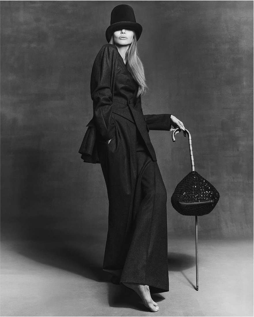 A woman posing in a dark suit with a hat and bare feet, holding an umbrella-like object, stands against a dark background in a studio shot.