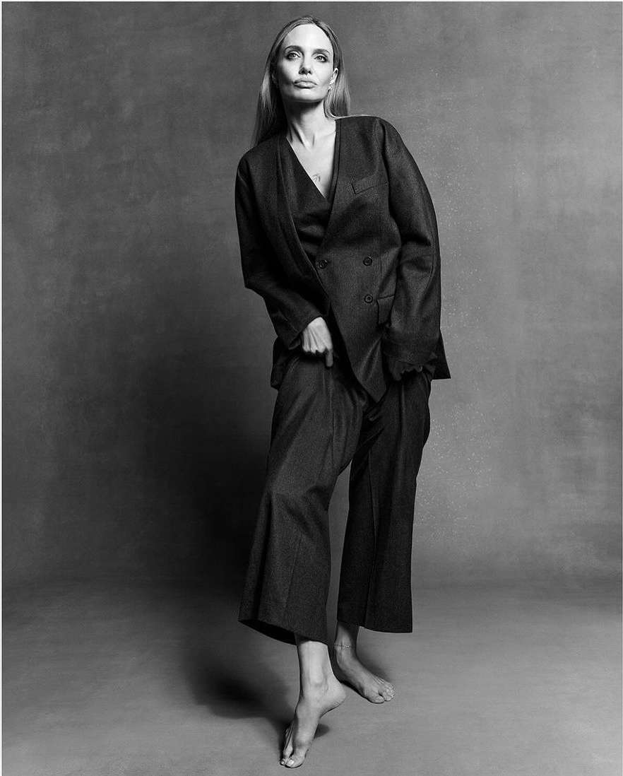 Black and white portrait of Angelina Jolie wearing a dark oversized suit against a textured grey background.